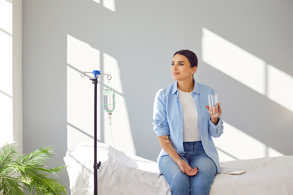 Woman receiving IV drip infusion therapy in her blood sitting in medical clinic with glass of water. Woman receiving IV drip infusion therapy in her blood sitting in medical clinic with glass of water.